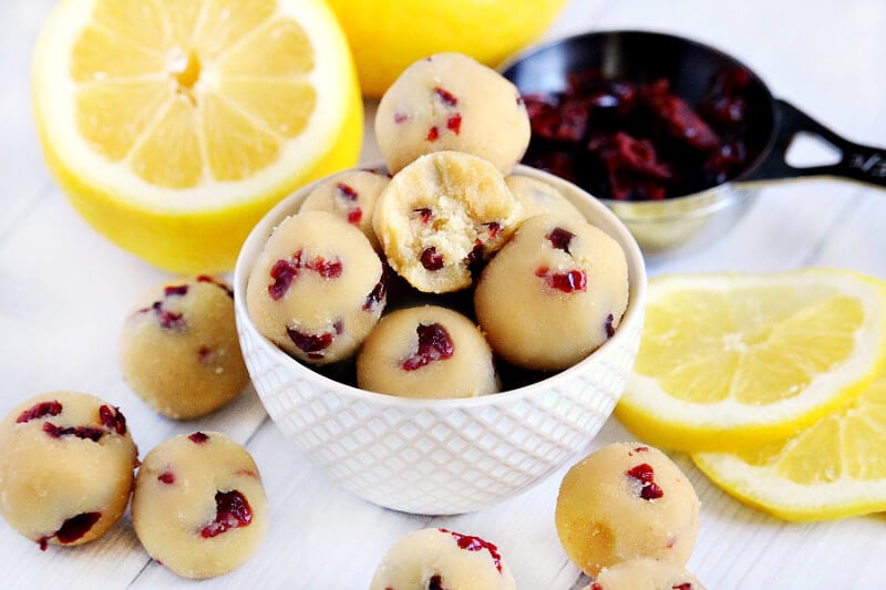 Horizontal image of Gluten-Free Vegan No-Bake Cranberry Lemon Ball Truffles in a cream colored bowl surrounded by lemons and cranberries