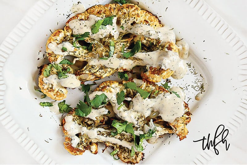 Horizontal image of Gluten-Free Vegan Roasted Cauliflower "Steaks" with Lemon Dill Tahini Dressing on a white plate on a white marble surface