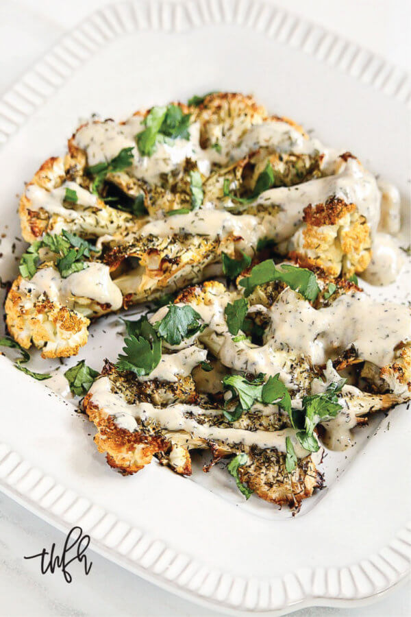 Vertical view of Gluten-Free Vegan Roasted Cauliflower "Steaks" with Lemon Dill Tahini Sauce on a white plate on a white marble surface