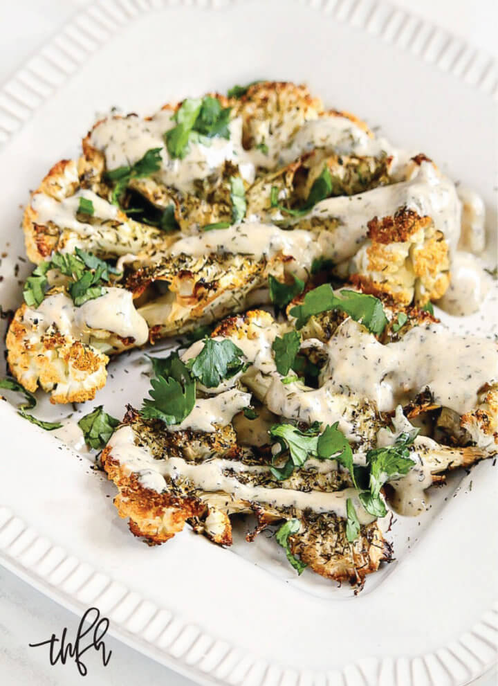 Vertical view of Gluten-Free Vegan Roasted Cauliflower "Steaks" with Lemon Dill Tahini Sauce on a white plate on a white marble surface