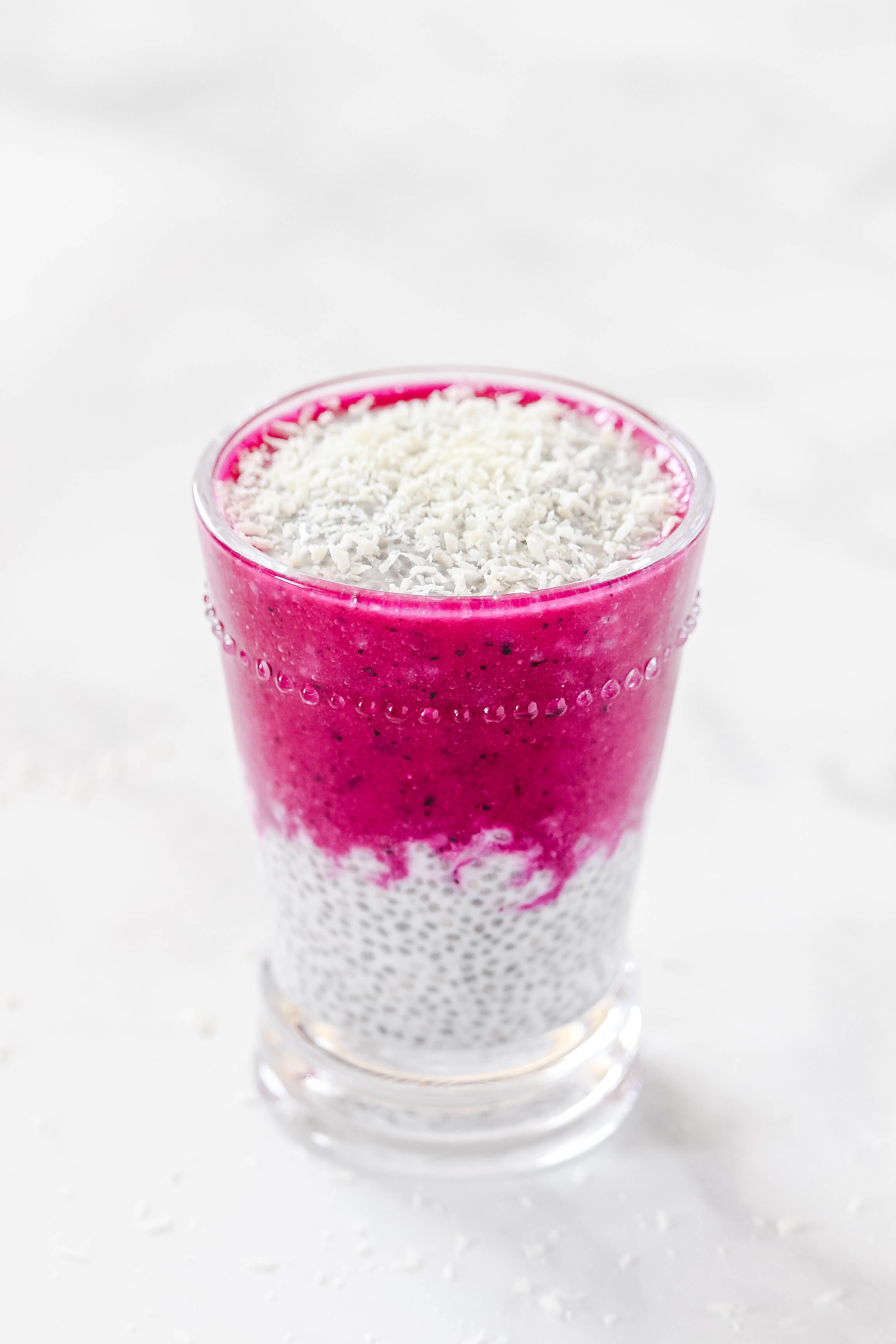 Vertical image of Gluten-Free Vegan Pitaya Dragon Fruit Chia Seed Pudding in a glass on a white marble surface
