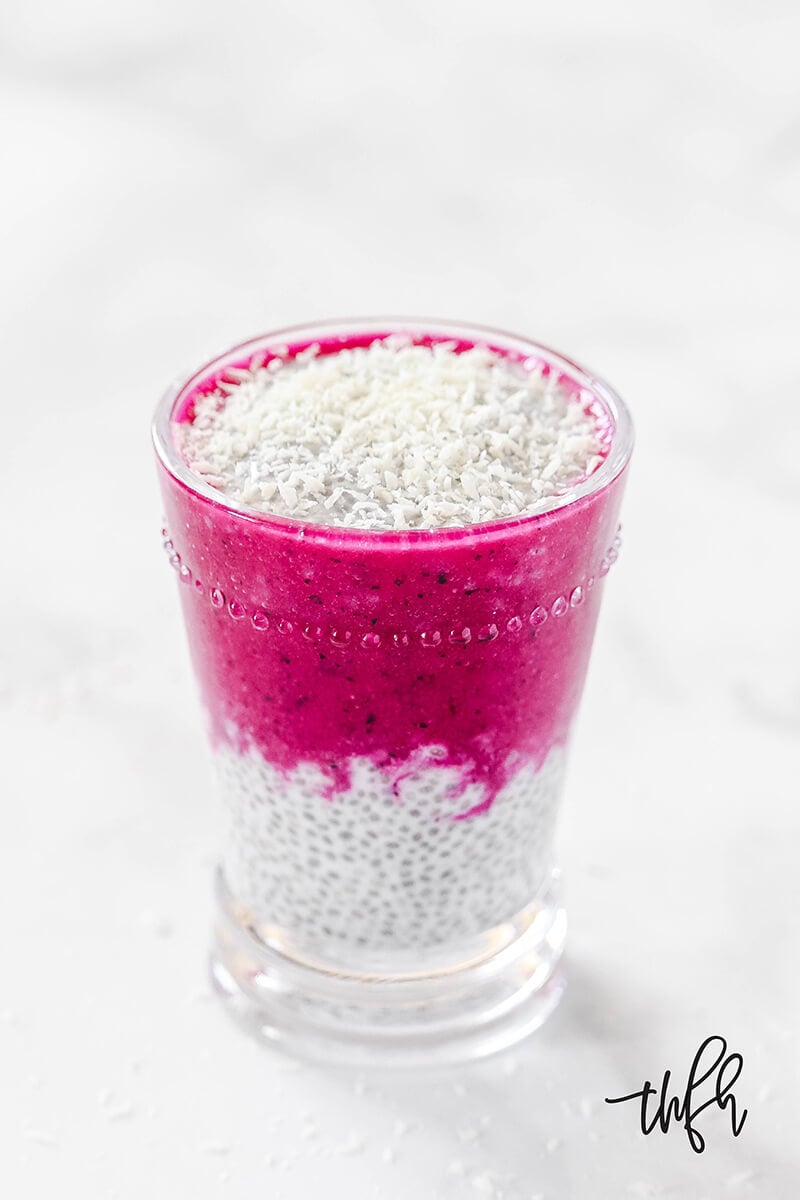 Vertical image of Gluten-Free Vegan Pitaya Dragon Fruit Chia Seed Pudding in a glass on a white marble surface