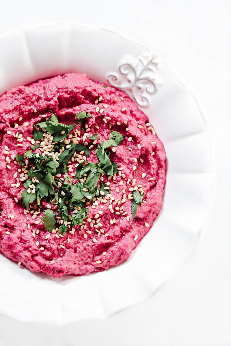 Overhead view of a decorative white bowl filled with The BEST Gluten-Free Vegan Spicy Jalapeno Beet Hummus on a solid white background