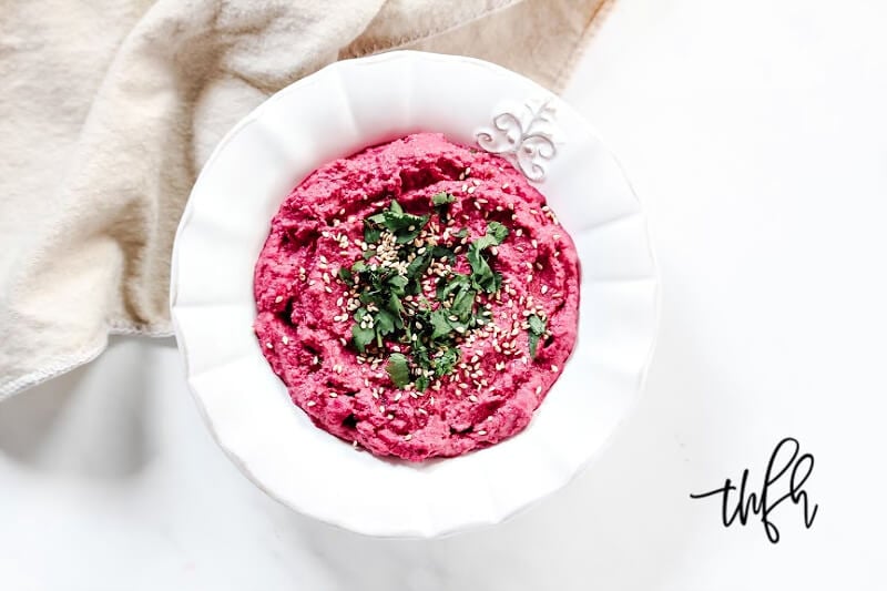 Overhead view of a white bowl of The BEST Gluten-Free Vegan Spicy Jalapeno Beet Hummus with a tan napkin in the background on a solid white surface