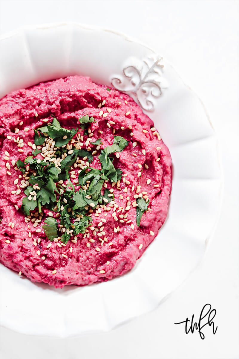 Overhead view of a decorative white bowl filled with The BEST Gluten-Free Vegan Spicy Jalapeno Beet Hummus on a solid white surface