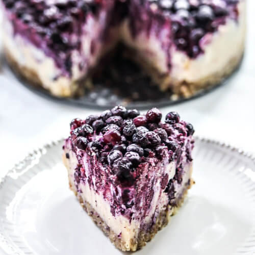 A single slice of Gluten-Free Vegan No-Bake Wild Blueberry Cheesecake on a small grey plate with the entire cheesecake in the background