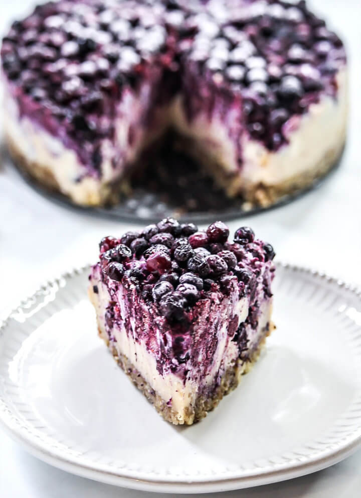 A single slice of Gluten-Free Vegan No-Bake Wild Blueberry Cheesecake on a small grey plate with the entire cheesecake in the background