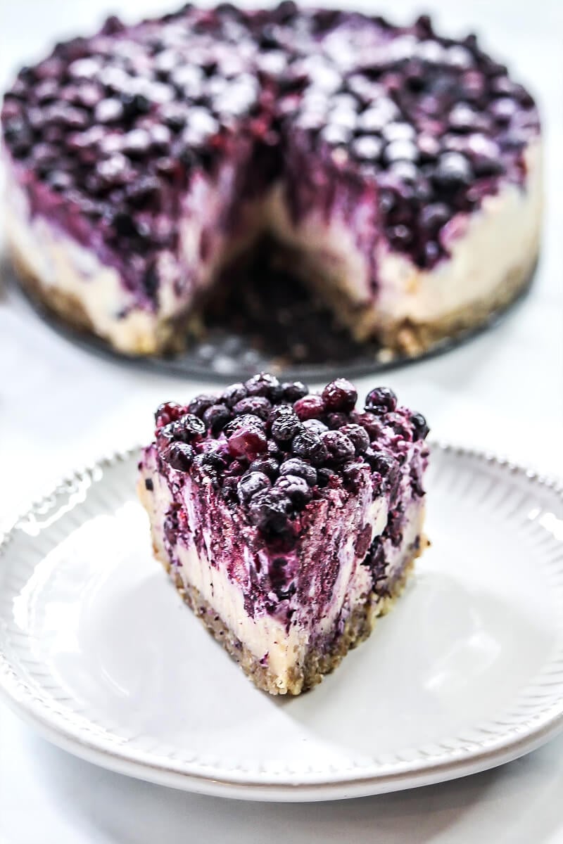 A single slice of Gluten-Free Vegan No-Bake Wild Blueberry Cheesecake on a small grey plate with the entire cheesecake in the background