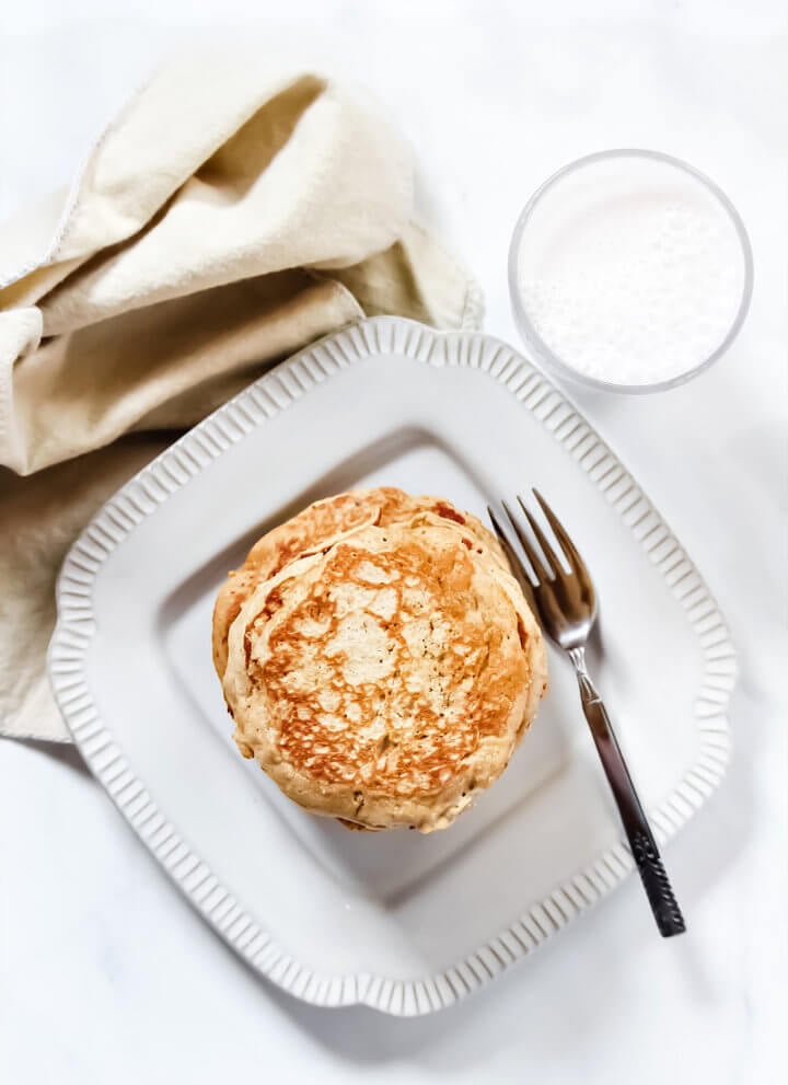 Overhead image of a decorative grey plate with a stack of Gluten-Free Vegan Pumpkin Spice Blender Pancakes on it with a glass of milk and cream cloth napkin to the side of the plate