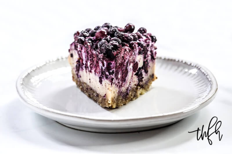 Head-on image of a single slice of Gluten-Free Vegan No-Bake Wild Blueberry Cheesecake on a small grey saucer on a white background