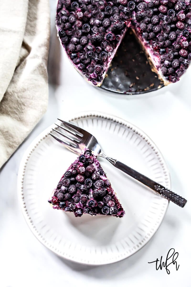 Overhead image of a small grey plate with a slice of Gluten-Free Vegan No-Bake Wild Blueberry Cheesecake next to a cheesecake pan with the remaining cheesecake