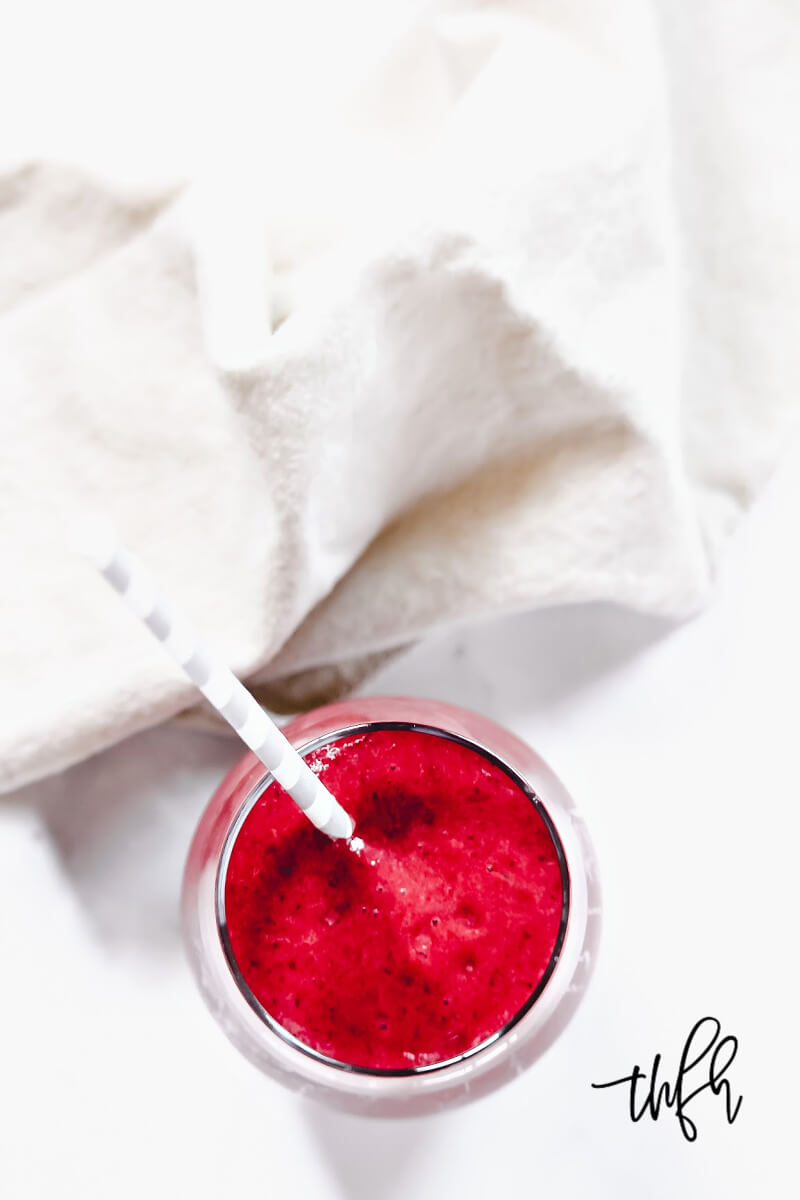 Overhead image of the top of a glass filled with The BEST Vegan Cherry Banana Smoothie with a grey and white straw and cream napkin beside the glass