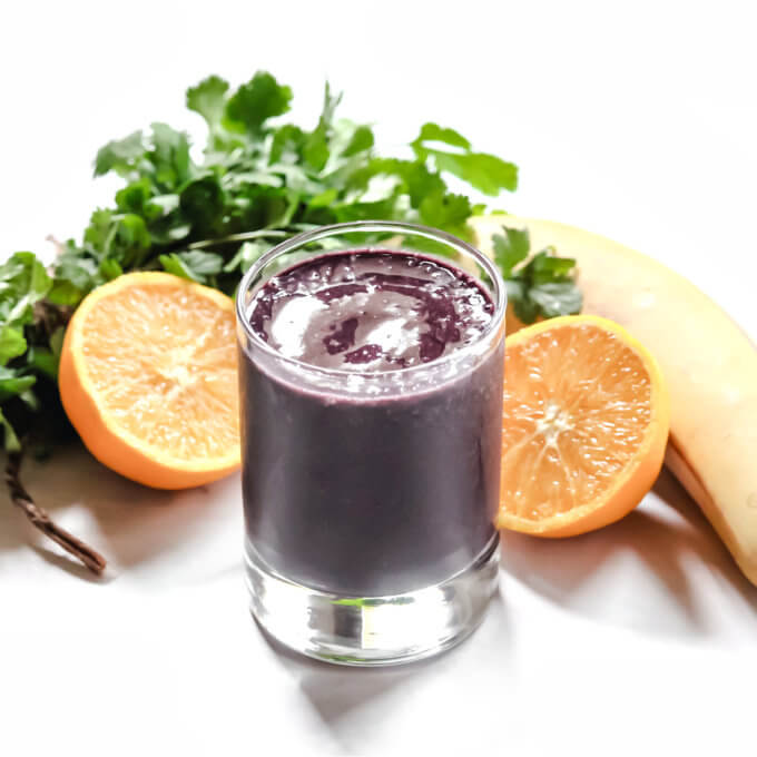 Small glass of dark purple smoothie surrounded by various fruits and veggies on a white background