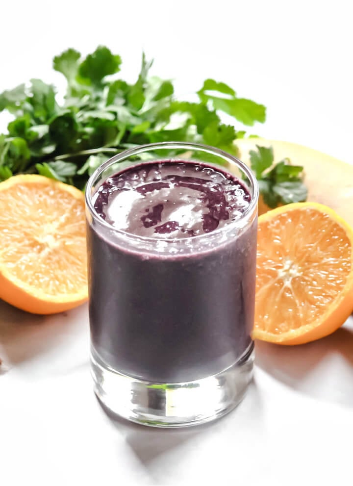 Small glass of dark purple smoothie surrounded by various fruits and veggies on a white background