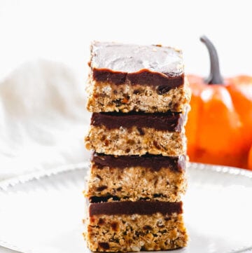 A stack of four Gluten-Free Vegan No-Bake Crispy Pumpkin Spice Squares in the center of a white plate with a small pumpkin in the background