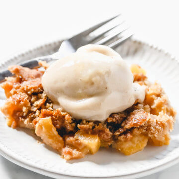 Vertical image of a grey plate filled with peach cobbler with a scoop of vanilla ice cream on top on a solid white surface