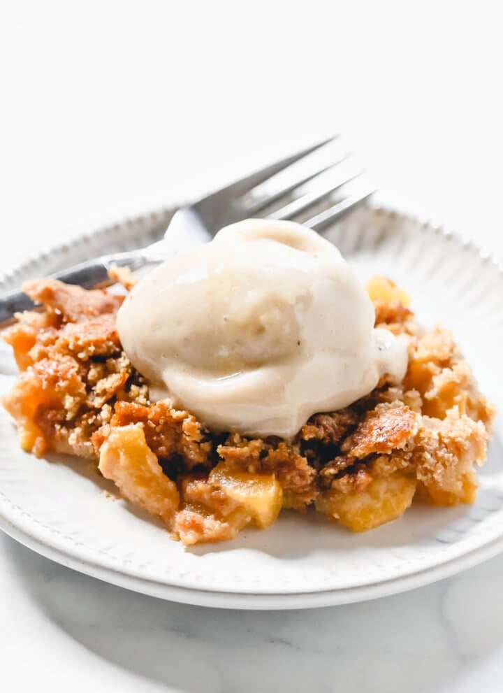 Vertical image of a grey plate filled with peach cobbler with a scoop of vanilla ice cream on top on a solid white surface