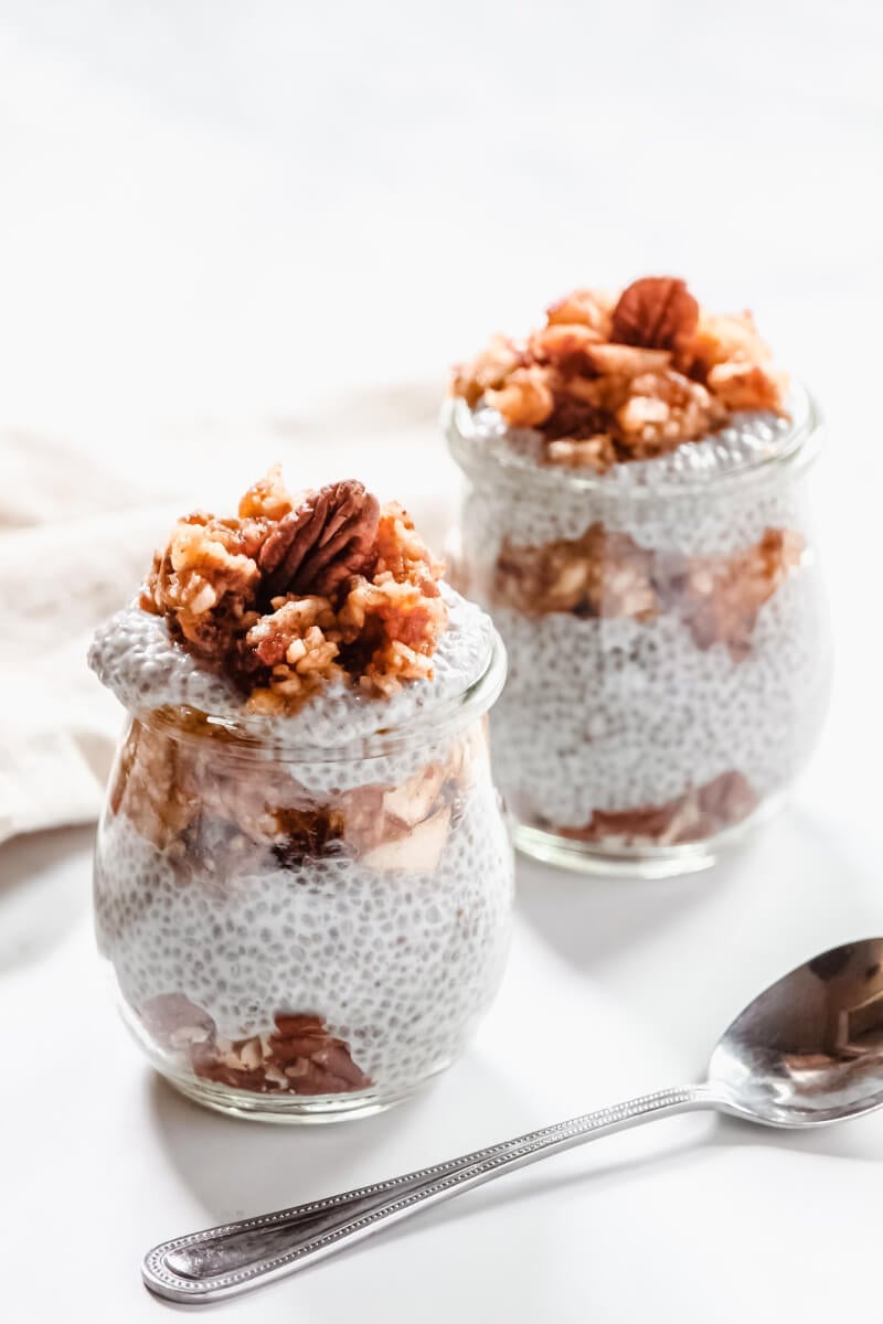 Two small glasses side-by-side filled with Gluten-Free Vegan Apple Pie Chia Seed Pudding on a white surface next to a silver spoon
