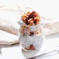 Horizontal image of a small glass of Gluten-Free Vegan Apple Pie Chia Seed Pudding on a white surface next to a silver spoon and cream colored napkin