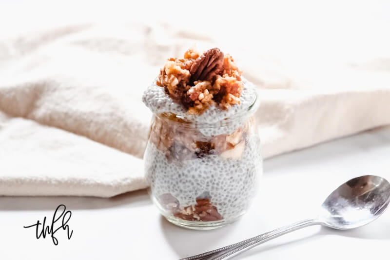 Horizontal image of a small glass filled with Gluten-Free Vegan Apple Pie Chia Seed Pudding on a white surface next to a silver spoon and cream cloth napkin