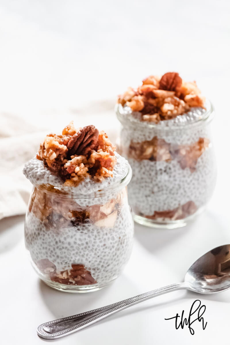 Two decorative glasses filled with Gluten-Free Vegan Apple Pie Chia Seed Pudding side-by-side on a white surface next to a silver spoon