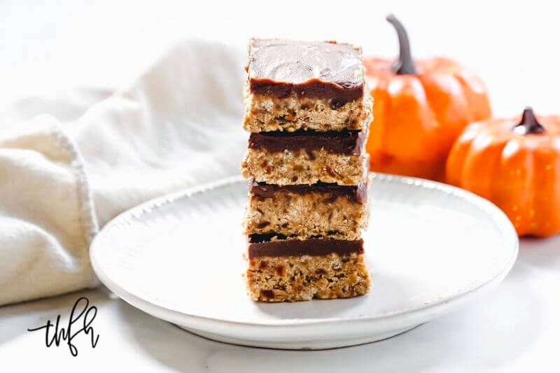 A stack of four crispy pumpkin bars covered with a layer of chocolate on a grey plate with two small pumpkins in the background
