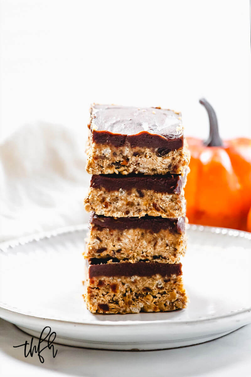 A stack of four Gluten-Free Vegan No-Bake Crispy Pumpkin Spice Squares in the center of a white place with a small pumpkin in the background