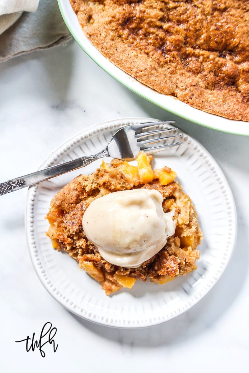 Overhead image of a plate filled with peach cobbler and a scoop of ice cream on top with a baking dish in the background