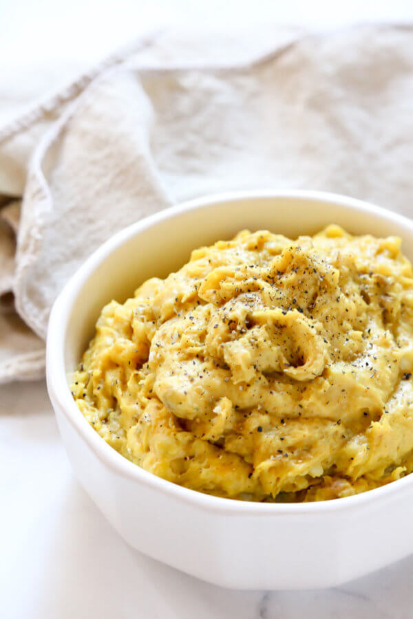 Close-up vertical image of a white bowl filled with Gluten-Free Vegan Turmeric and Black Pepper Mashed Potatoes on a white marble surface with a cream cloth napkin next to it