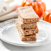 Horizontal image of a stack of four Gluten-Free Vegan No-Bake Pumpkin Spice Granola Bars on a decorative grey plate with two small pumpkins in the background