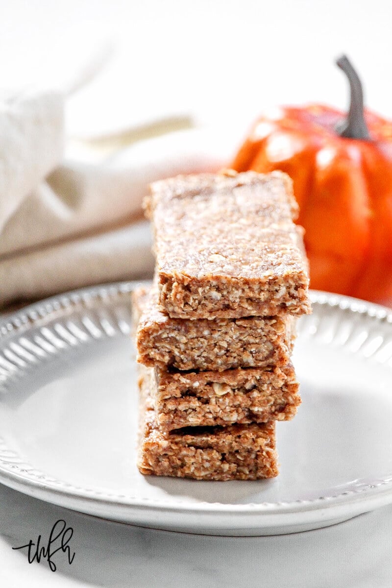 Vertical image of a stack of four Gluten-Free Vegan No-Bake Pumpkin Spice Granola Bars on a small grey plate with a cloth napkin and small pumpkins in the background