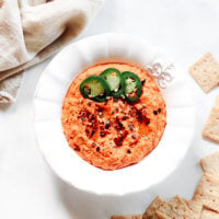 Overhead vertical image of a decorative white bowl filled with red pepper hummus next to crackers and a cream cloth napkin