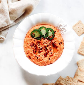 Overhead vertical image of a decorative white bowl filled with red pepper hummus next to crackers and a cream cloth napkin