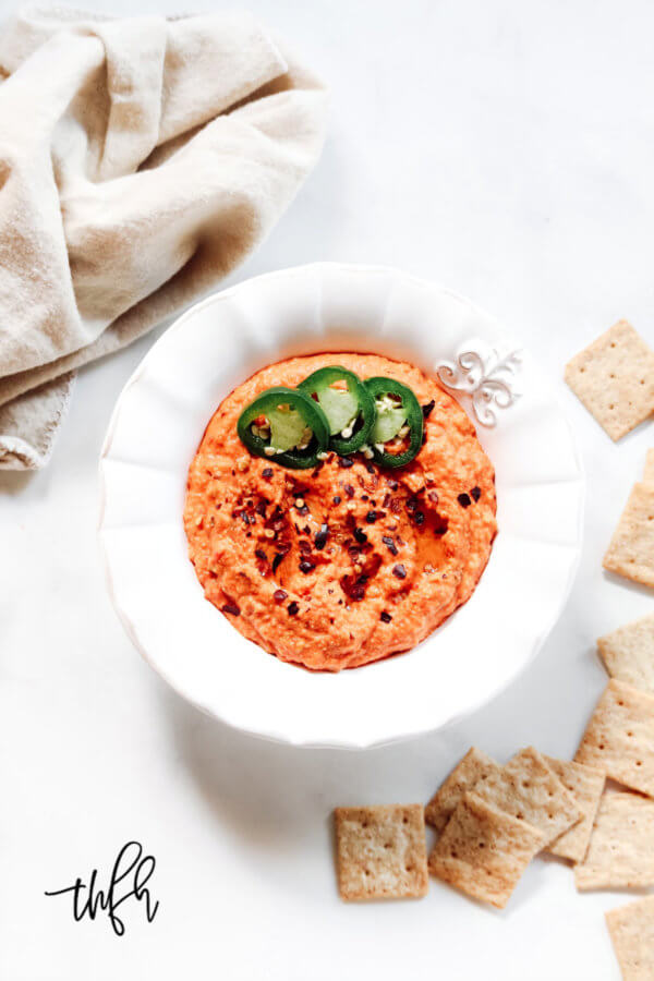 Overhead vertical image of a decorative white bowl filled with red pepper hummus next to crackers and a cream cloth napkin