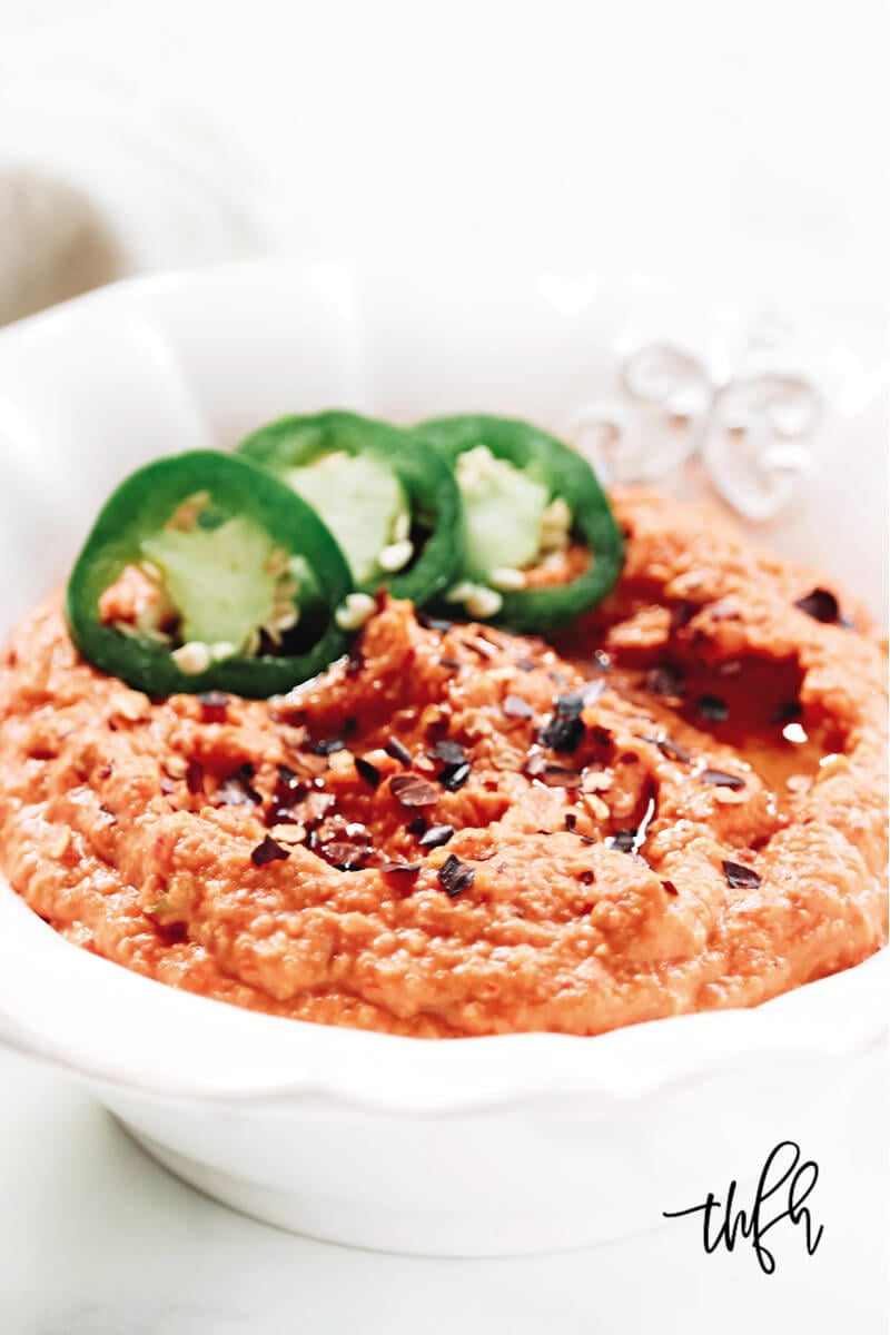 Vertical image of a white decorative bowl filled with Gluten-Free Vegan Spicy Roasted Red Pepper and Garlic Hummus garnished with fresh jalapenos and red pepper flakes
