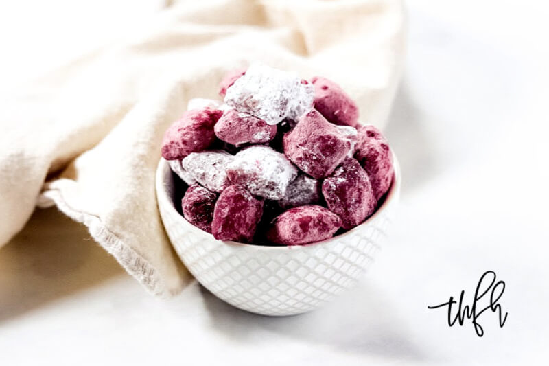 Horizontal image of a small decorative white bowl filled with red and white powdered sugar puppy chow treats on a solid white surface with a cream colored cloth napkin in the background