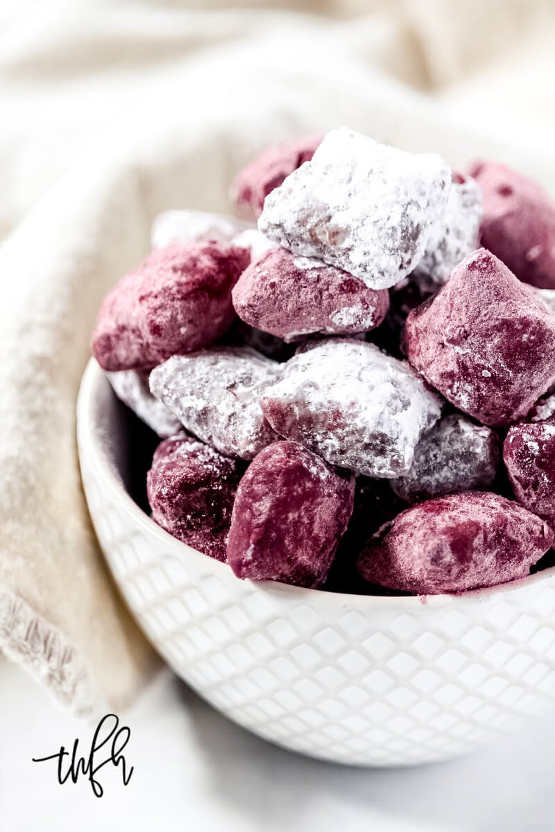 Vertical image of a small decorative white bowl filled with red and white powdered pieces of puppy chow with a cream cloth napkin in the background