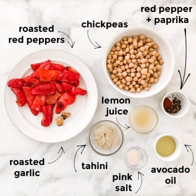 Flat-lay of ingredients needed to make roasted red pepper hummus labeled on white marble surface