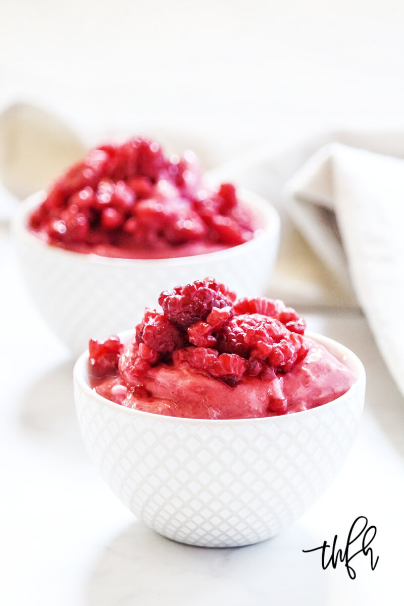Two small decorative white bowls filled with Gluten-Free Vegan Raspberry Banana "Nice" Cream on a white marbled surface with a cream cloth napkin to the side