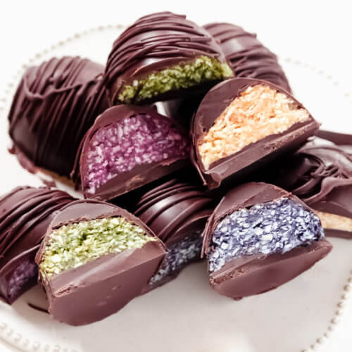 Vertical image of multiple chocolate covered colored coconut eggs on a decorative white platter on a solid white background