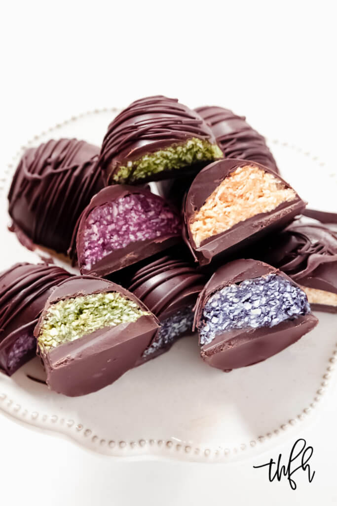 Vertical image of multiple chocolate covered colored coconut eggs on a decorative white platter on a solid white background