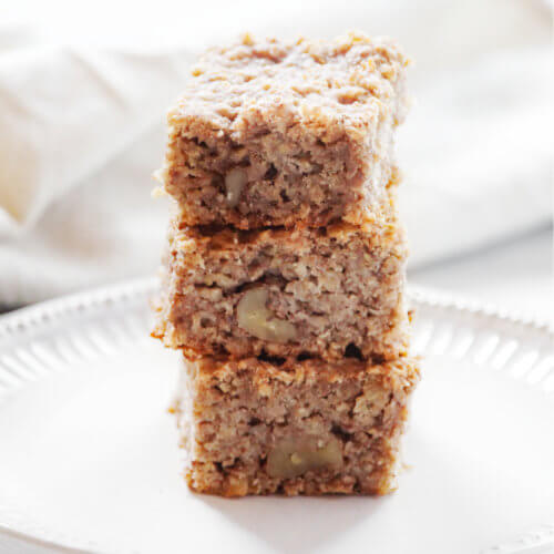Square image of a stack of three baked oatmeal squares on a decorative white plate with a cream cloth napkin in the background