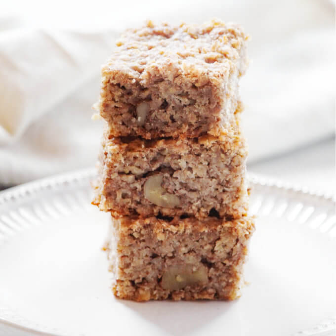Square image of a stack of three baked oatmeal squares on a decorative white plate with a cream cloth napkin in the background
