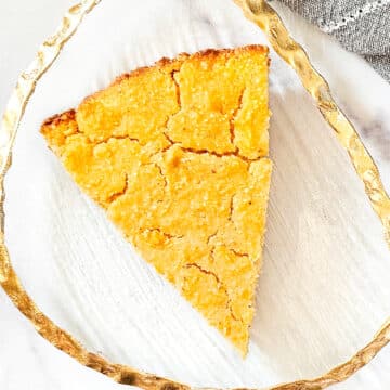 Overhead image of a slice of vegan skillet cornbread on a gold trimmed glass plate next to a grey napkin on a white marble surface.