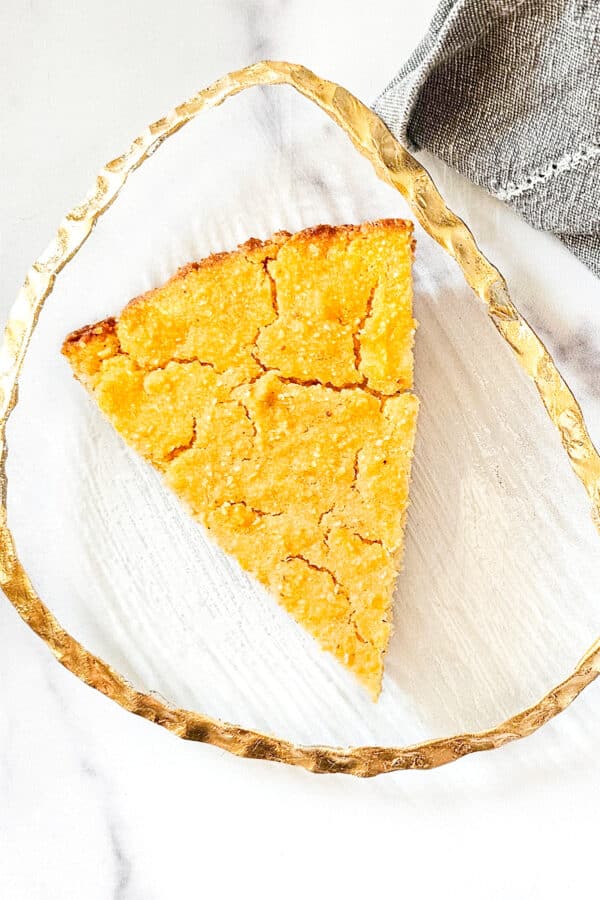 Overhead image of a slice of vegan skillet cornbread on a gold trimmed glass plate next to a grey napkin on a white marble surface.