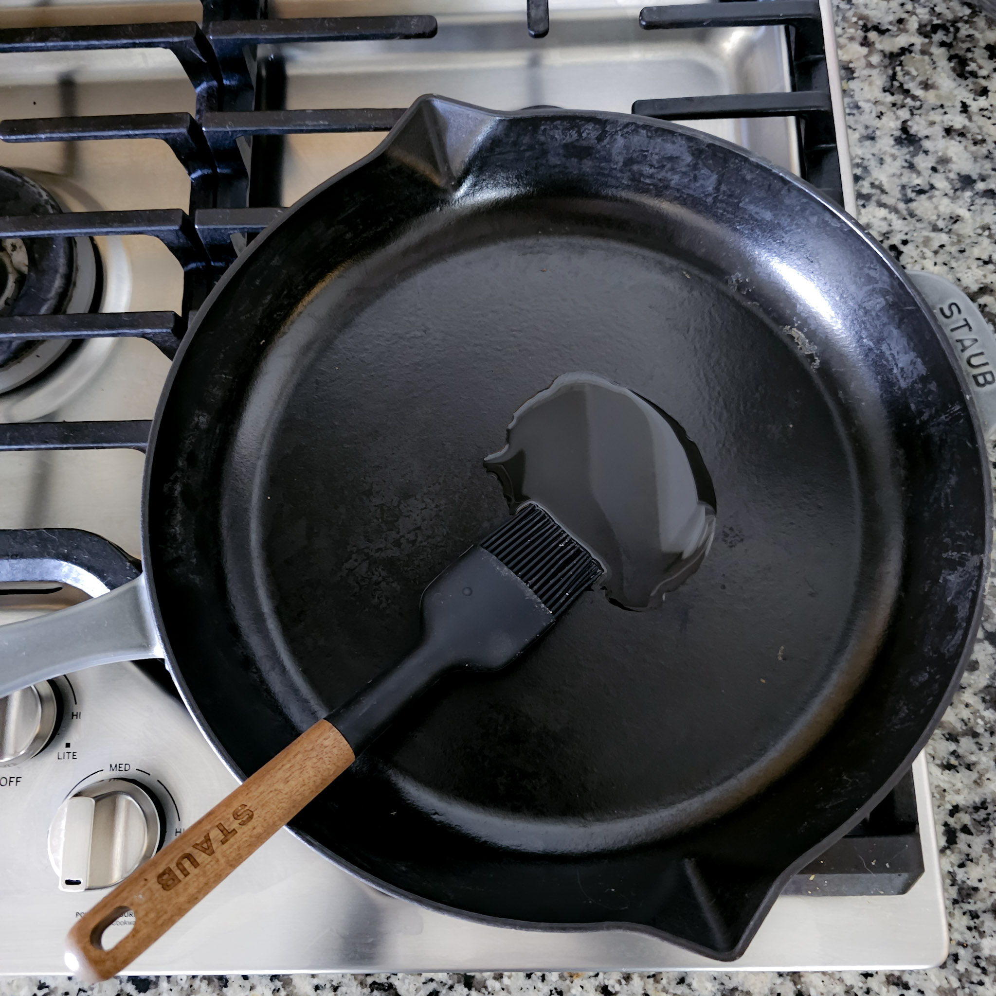 Overhead image of avocado oil being spread evenly onto a cast iron skillet over a gas cooktop with a wooden utensil.