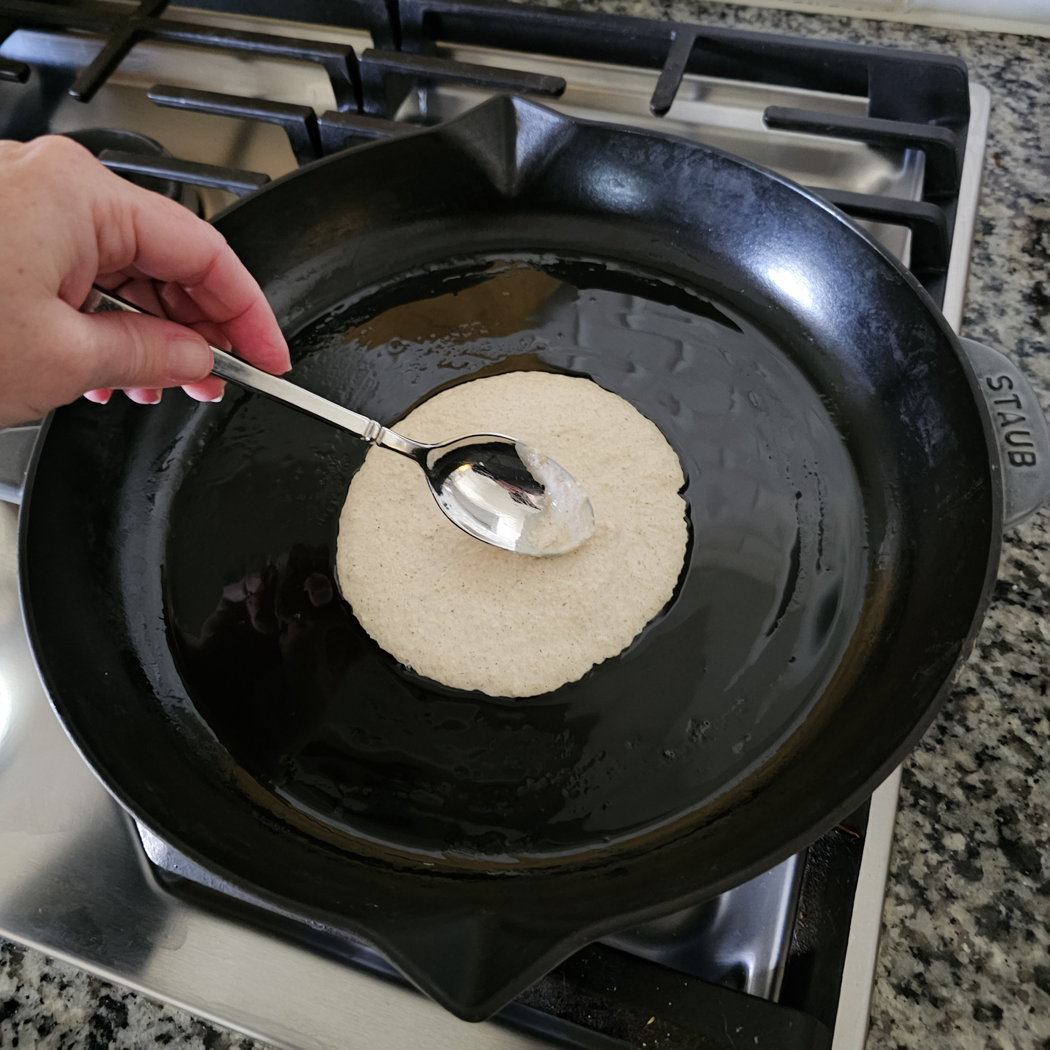 Overhead image of mixture to make gluten-free vegan oat tortillas being spread evenly on a cast iron skillet with a spoon on top of a gas cooktop.
