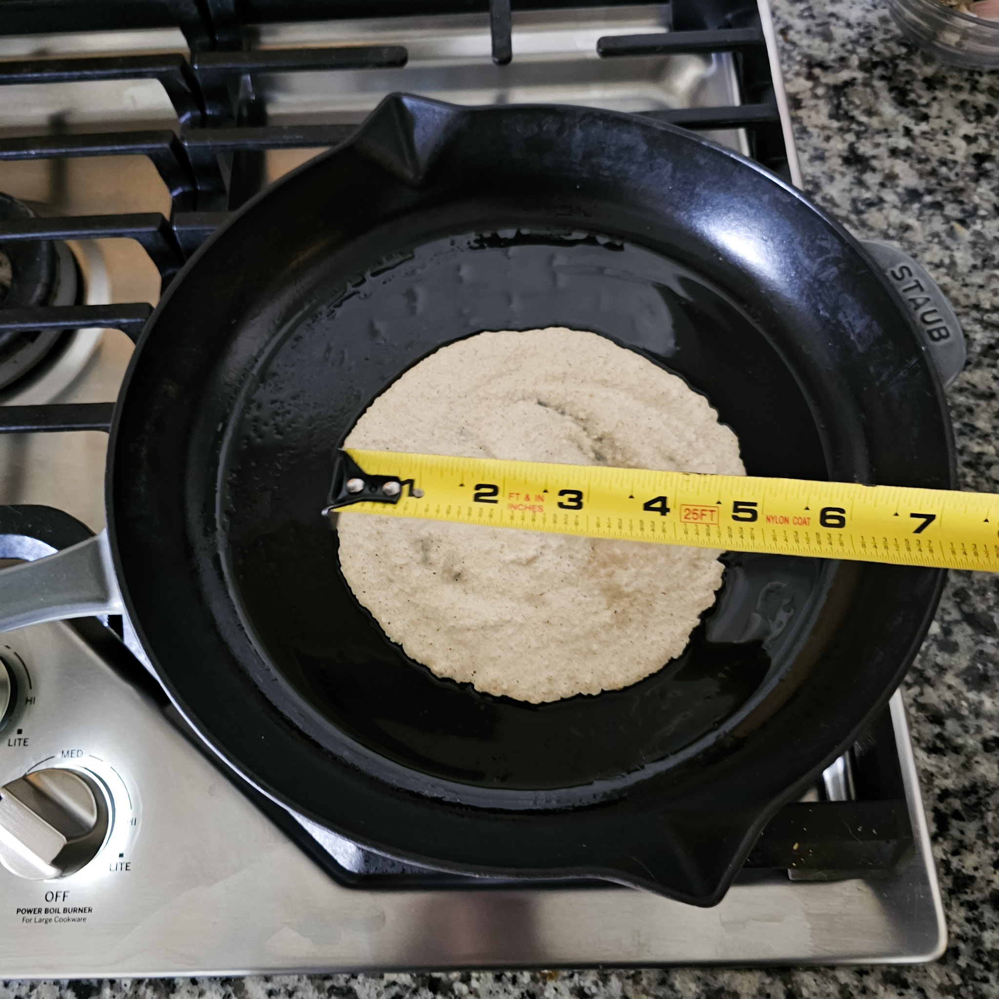 Overhead image showing a tape measure measuring 5 inches over a gluten-free vegan oat tortilla in a cast iron skillet over a gas cooktop.