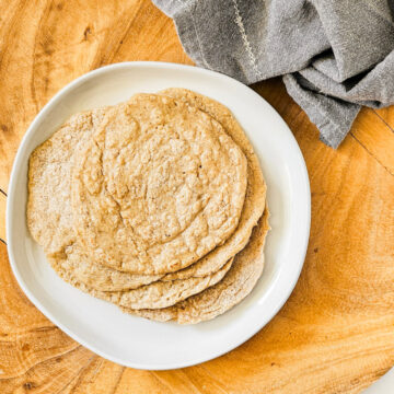 Overhead image of a stack of gluten-free vegan oat tortillas on a white plate on a wooden surface next to a grey linen napkin.