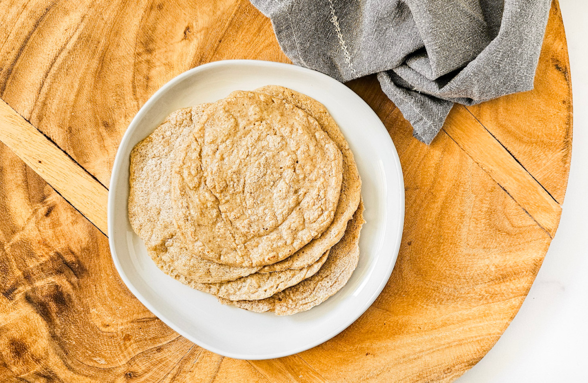 Overhead image of a stack of gluten-free vegan oat tortillas on a white plate on a wooden surface next to a grey linen napkin.